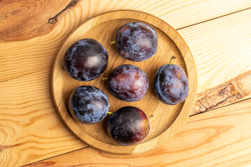 Several organic black plums on a round wooden tray, close-up, on a wooden table, top view.