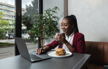 Young happy college exchange student or African American freelancer business woman having issue eating big fat tasty cheeseburger with melting cheese while she work on laptop computer in cafeteria. 