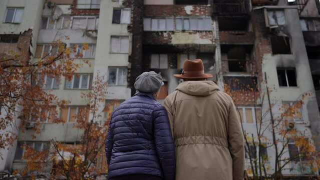 Back view senior couple looking at ruined residential building talking standing outdoors in city in Ukraine. Old Caucasian man and woman discussing lost property. Russian-Ukrainian war concept