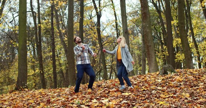 Funny young couple dancing in forest with trees and leaves in autumn having fun listening to music. Beautiful blonde stylish happy woman and bearded attractive man spending time together moving crazy.