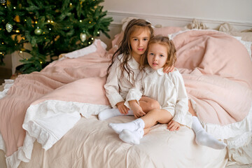 Fototapeta na wymiar Two little curly-haired sisters are sitting on the bed in their nightgowns on the morning after Christmas. Soft pink bedding on the bed