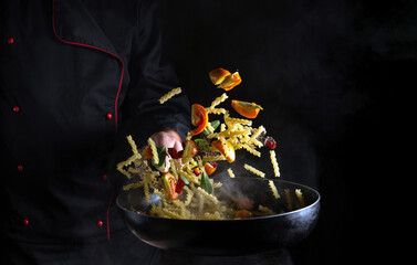 The cook prepares and throws food on a hot pan with vermicelli with vegetables. Place for advertising on a black background