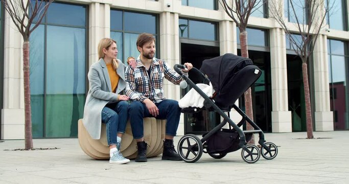 Young happy family sitting enjoying fresh air. Attractive loving blonde young mom hugging handsome caring bearded adult dad looking at newborn baby in stroller smiling. Parenthood concept.