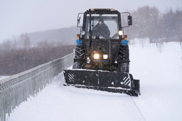 The work of the municipal snow removal service. A wheeled tractor counts snow from a pedestrian path as a front snow dump. Through the snowy haze, the road is illuminated by headlights.