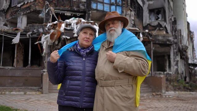 Medium shot portrait of sad Ukrainian senior couple with national flag in shoulders posing in bombed destroyed city. Frustrated man and woman looking at camera standing outdoors. Slow motion