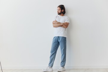 A man with a beard and long hair in a white T-shirt and blue jeans stands against a white wall...