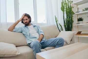 A young male freelancer sits on the couch after a demanding job and rests sadly leaning on the couch. 