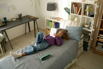High angle view of little girl lying on her bed and reading her diary during her leisure time at...