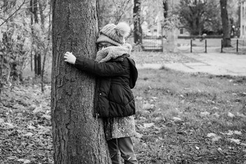 Dziewczynka przytula się do drzewa w parku, kocha przyrodę, jesienny spacer popołudniowy, dziecko w kurtce, czapce i szalu, zima, zimowy spacer - obrazy, fototapety, plakaty