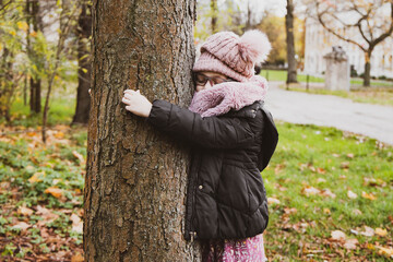Rozmarzona dziewczynka przytula się do drzewa, dziecko na dworze, w parku, kurtka i czapka, jesień, zima