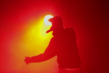 Silhouette of rap singer with microphone. Cool rapper with mic in hand singing on concert stage in bright red lights. Hip hop artist performing live on scene in music hall - obrazy, fototapety, plakaty