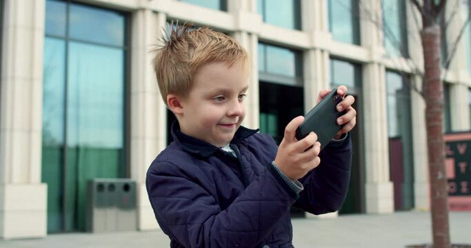 Close-up portrait of cute little happy boy standing outdoors in city playing racing games with accelerometer smiling laughing. Cheerful joyful child having fun using smartphone on fresh air.