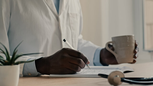 Male doctor in medical coat sit at clinic hold pen write prescription in notebook african man professional medic general practitioner physician make notes planning work day drinking tea coffee water