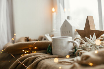 Warm cup of tea, christmas decorations, lights, little house, star on cozy blanket on windowsill. Winter hygge, Christmas still life. Cozy home on snowy day. Atmospheric scandinavian mood