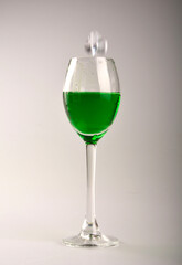 zielony płyn w kieliszku do wina, zielony napój w kieliszku na stole, zielony napój z kostkami lodu w kieliszku, absynt w kieliszku, wpadajaca do napoju kostka lodu, ruch, Kostka lodu wpada do drinka