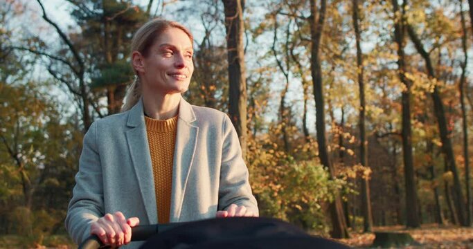 Close-up portrait of caring loving young mom walking with newborn kid in stroller enjoying sunny autumn day in park. Pretty blonde adult woman strolling outside with little child. Motherhood concept.