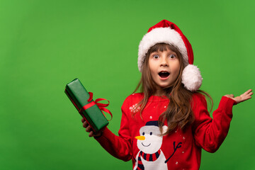 Portrait of an emotional surprised girl in a red knitted sweater and a Santa hat holds a beautiful gift in her hand. Surprise delight. Merry Christmas and Happy New Year 2023