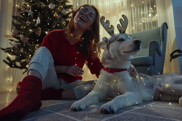 Young woman on Christmas evening together with pet dog having fun putting on Christmas deer horns and garland sitting on the floor to the Christmas tree