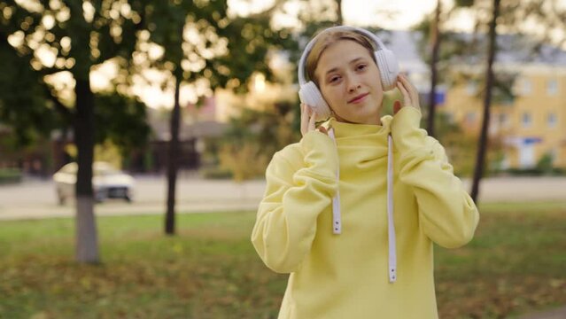 Young girl walking along city street, lifestyle. Girl goes to park in autumn, listens to music in headphones. Young active woman walks outdoors wears headphones, listens to audio books, has rest.