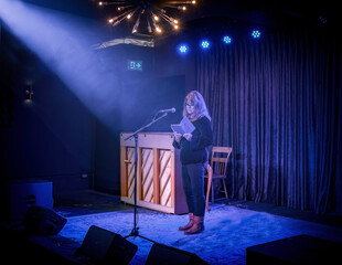 Poet Deborah-Anne Tunney reading from A Different Wolf on a nightclub stage, stage lights, spot lights, piano, microphone and microphone stand