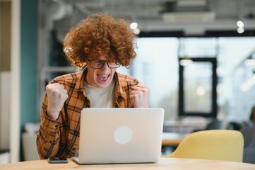 Young happy male freelancer in casual clothes sitting in cafe with laptop and using mobile phone.