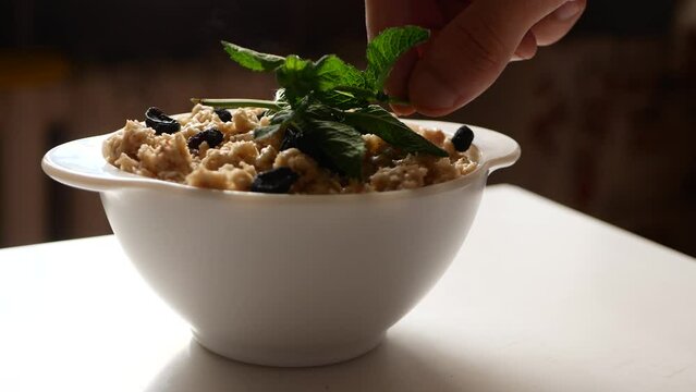 Sprigs Of Green Mint Are Placed On Plate Of Steaming Oatmeal With Black Raisins.