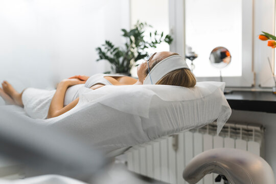 Back indoor view of unrecognizable caucasian woman in white tank-top and protective hairband lying comfortably on SPA bed and waiting for her facial treatment. High quality photo