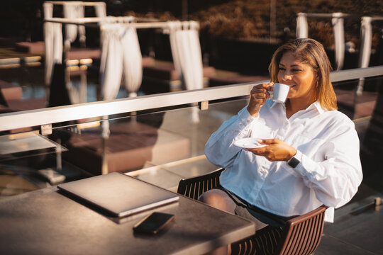 A dazzling plus-size Hispanic female entrepreneur drinking a coffee while sitting at the table with a laptop and smartphone in an outdoor cafe, enjoying golden hour after finishing a freelance project