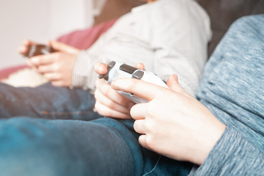 Cropped photo of two teenage boys children sitting on sofa at home, holding gaming controller joystick gamepad, playing videogames on game console. Hobby, free time, gaming, entertainment, leisure.