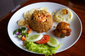 Javanese fried rice with egg and fried chicken on a white wide plate. There are additional slices of cucumber, lettuce, tomatoes and emping crackers.