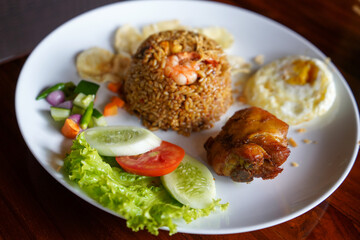 Javanese fried rice with egg and fried chicken on a white wide plate. There are additional slices of cucumber, lettuce, tomatoes and emping crackers.