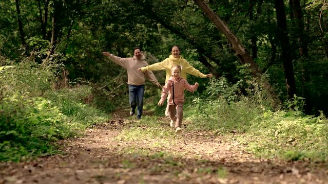 Mom, dad, daughter play in park, run along path together. Happy child run away from parents. Happy family walks in forest, spring, summer. Teamwork. Weekend with family. Concept of childrens happiness