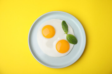 Tasty fried eggs with basil in plate on yellow background, top view