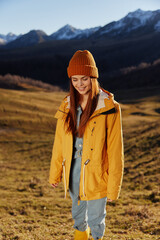 Woman running up the hill to the camera smile with teeth in the mountains in the autumn in a yellow raincoat and jeans happy sunset trip on a hike mountains in the snow, freedom lifestyle 