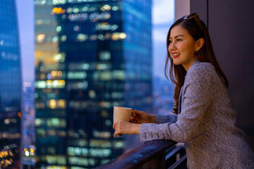 Young Asian woman enjoying the evening view from her balcony while looking at the urban skyscraper...