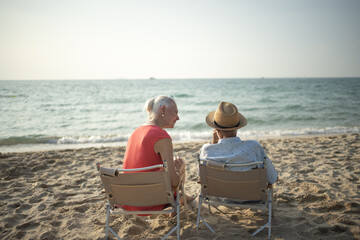 Two elderly men and women sit chair at the beach talking and watching the sun and the sea on their summer vacation and they smile and enjoy their vacation.