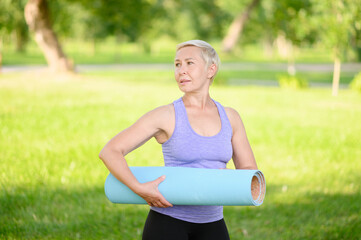 Middle-aged woman doing smiling and holding a fitness mat outside, leading a healthy lifestyle.Time to sport.