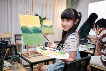 Portrait of Asian girl looking at camera and smiling with acrylic color picture painted on canvas in art classroom and creative learning with talents and skill at elementary school studio education.