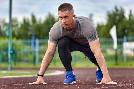 Serious Caucasian athlete trains in a stadium with music in wireless headphones. Healthy young man prepared for a training run around the stadium in the open air.