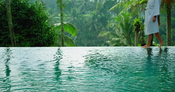 Amazing rice terraces near to swimming pool in sunny morning Bali with woman silhouette walking in luxury tourist resort. Girl in bathrobe by poolside, palm forest jungle on tropical island.