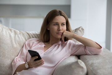 Pensive attractive woman rests seated on sofa at home with mobile phone in hand, feels concerned,...