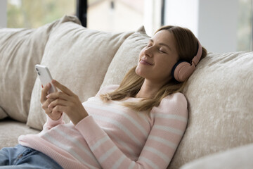 Serene young woman enjoy favourite music via wireless headphones, lying on sofa with smartphone,...
