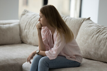 Obraz premium Thoughtful upset young 30s woman sits on sofa at home looks deep in thoughts, thinking about life concerns or break up feels unhappy, goes through divorce or marriage split, makes difficult decision