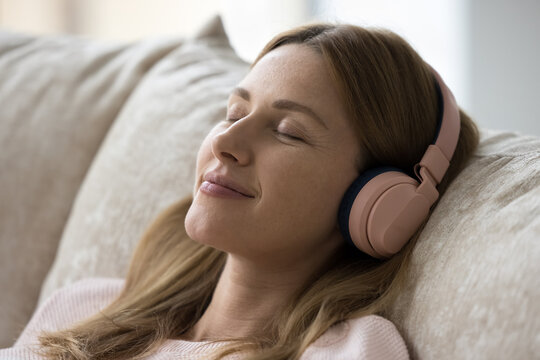 Close up face of peaceful beautiful woman listens music through wireless headphones relaxing on cushions of cozy couch. Digital streaming on-line services user, modern tech, weekend leisure and hobby