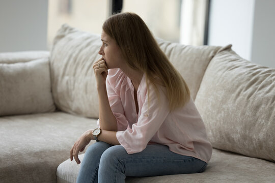Thoughtful upset young 30s woman sits on sofa at home looks deep in thoughts, thinking about life concerns or break up feels unhappy, goes through divorce or marriage split, makes difficult decision