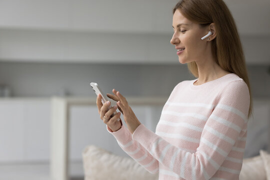 Attractive young woman standing in living room wear wireless earphones looks at smartphone screen smile choose music, play favourite song, listen podcast spend free time at home use modern technology