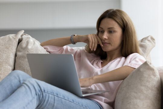 Serious pensive young woman sit leaned on sofa with laptop, do e-shopping at home, consider commercial offer, busy in telework task, thinks over ideas work indoor. Modern tech for business or leisure
