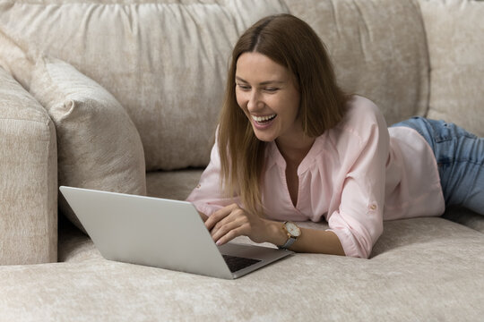 Smiling woman relax on sofa with laptop, watch on-line movie spend time at home on internet, staring at device screen enjoy new video vlog content looks happy. Modern tech, e-commerce services, fun