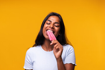 Young Indian woman eating pink popsicle on yellow background. Asian woman eating ice cream. - obrazy, fototapety, plakaty