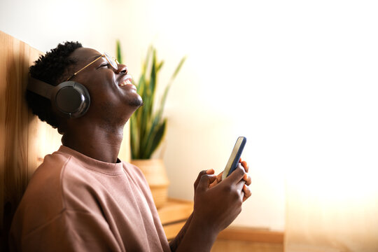 Young black man Listening to music with wireless headphones using mobile phone at home. Blissful moments. Copy space.